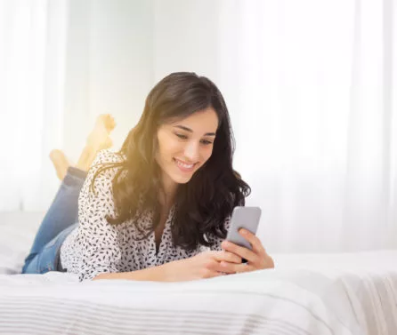 Smiling,Woman,Looking,At,Mobile,Phone,Lying,Down,On,White